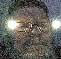 Close-up selfie of a man with glasses in low light.