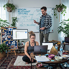 A satirical image of a couple, Megan and Tyler, attempting to manage an open relationship as a corporate startup. Tyler stands before a whiteboard labeled "Henson Relationship Ventures" covered in complex flowcharts and the phrase "Open to New Energy," while Megan sits on a rug with a laptop, looking distressed.