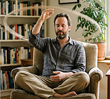 Man practicing hands on meditation. He sits in an armchair with eyes closed, making a sweeping lasso motion with his right hand above his head.