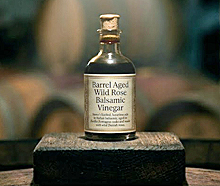 A close-up of a small glass bottle, labeled Noma Projects Barrel Aged Wild Rose Balsamic Vinegar, on a wooden block in a cellar with a spotlight.