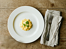 Overhead view of scrambled egg technique result: soft golden curds on white plate, copper pan, linen napkin, silver fork, rustic wood surface.