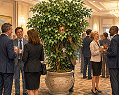 Editorial illustration about introversion showing a man hiding behind a large ficus at a busy conference while cheerful professionals network nearby.