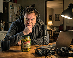 A man sits at a kitchen table staring angrily at a jar of pickles while podcast equipment waits nearby, moments before starting a podcast.